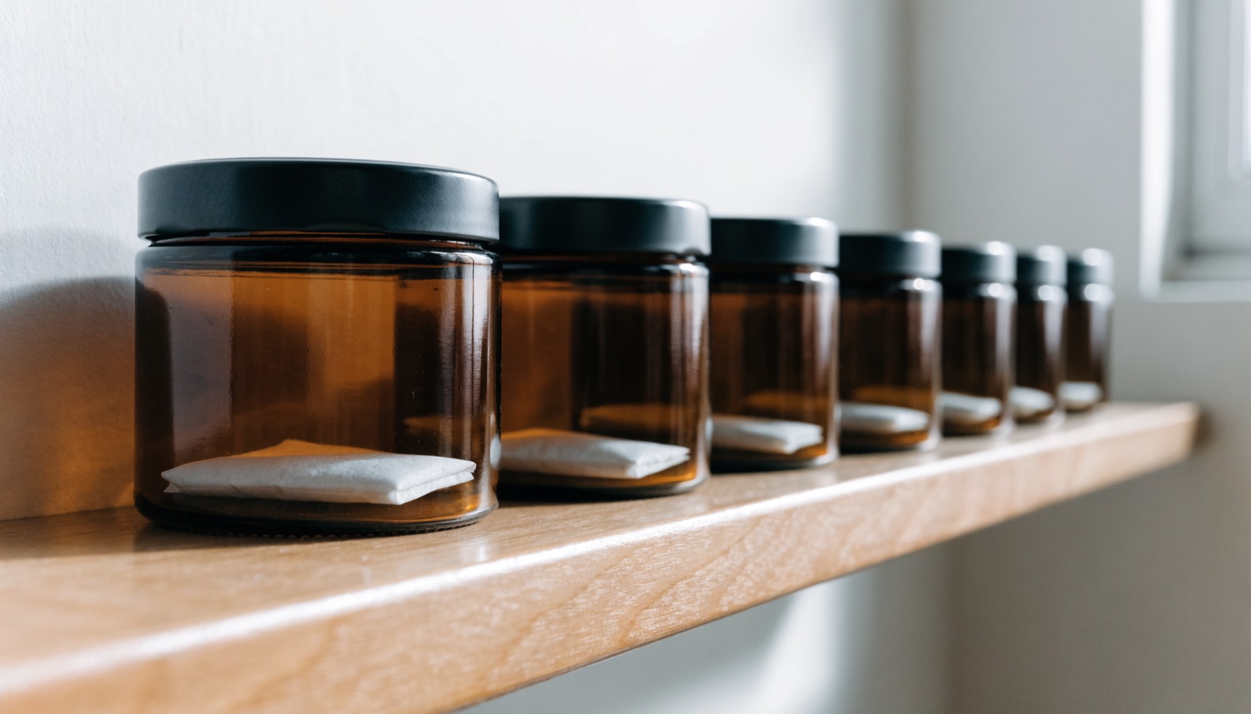 A row of identical amber glass jars with matte black lids on a pale oak shelf, each labelled as a Toad Venom lot and holding a small flat white humidity control sachet