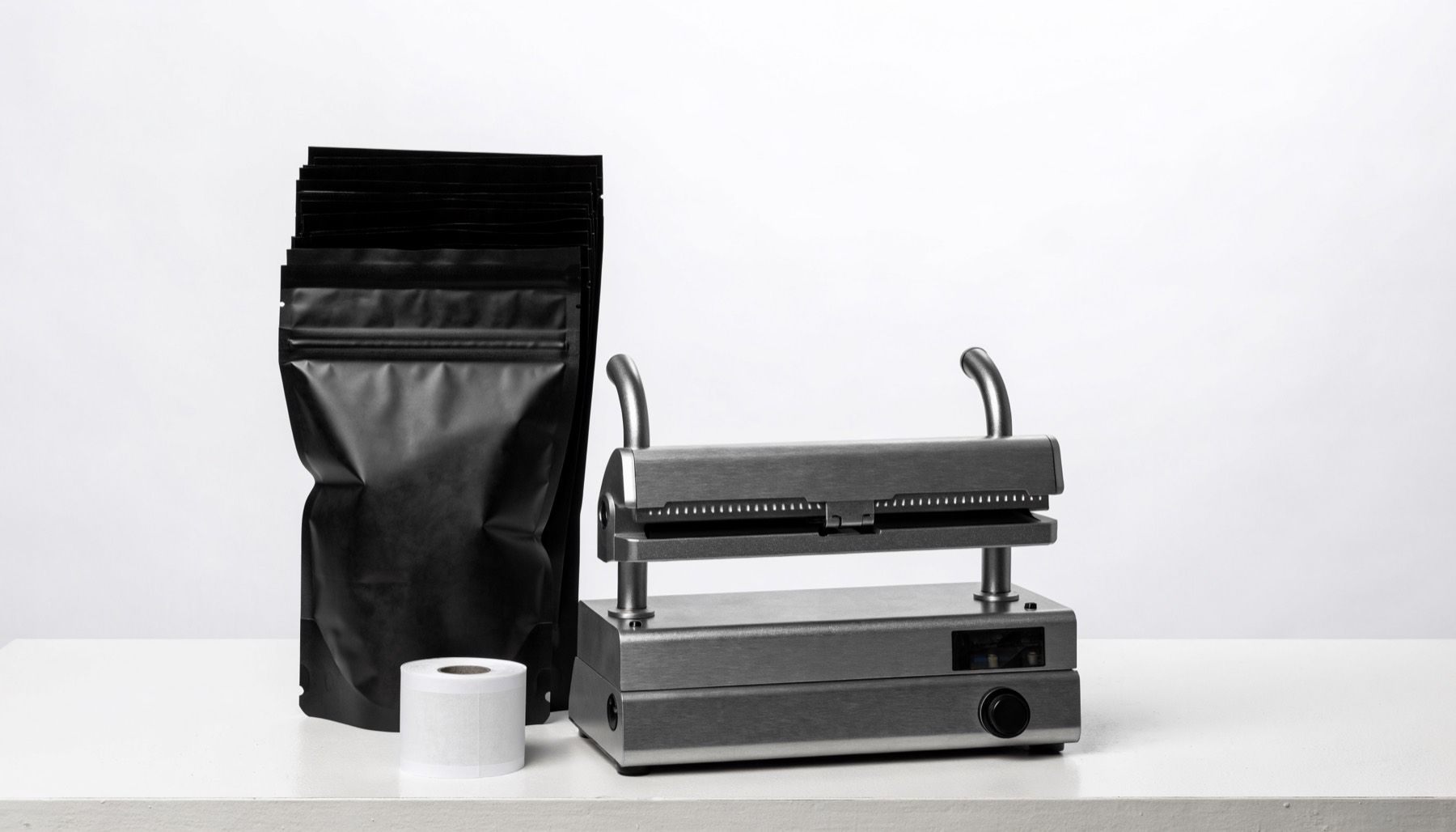 A tabletop impulse heat sealer on a white workbench beside a stack of matte black foil-lined stand-up pouches for an Uncle Snoop lot and a roll of blank white labels