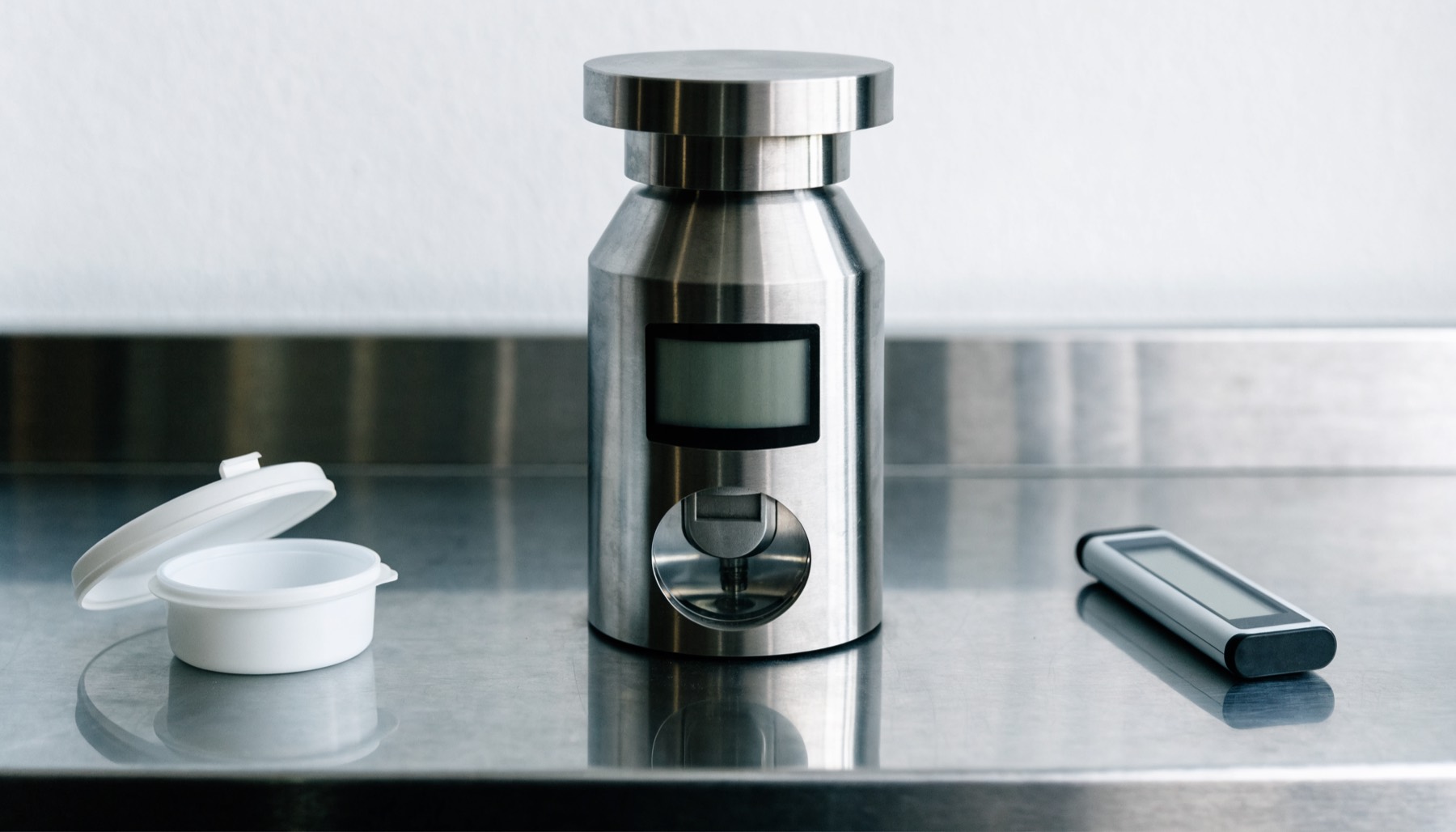 A benchtop water activity meter on a stainless steel bench beside an open white sample cup and a slim digital thermo-hygrometer probe